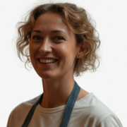 Julia Williams med kort krøllet hår smiler til kameraet, iført lysegrå trøje og blå remme over skuldrene mod hvid baggrund.
